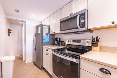 Bright modern galley kitchen with stainless steel refrigerator, over-the-range microwave and glass-top stove, white cabinets, beige countertops, coffee maker and knife block