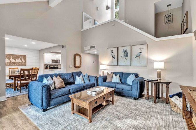 Open-plan coastal-inspired living room with high vaulted ceilings, two blue sofas with patterned pillows, wooden coffee table on a light-gray rug, framed heron artwork above, lamps on side tables, and a dining area and kitchen visible in the background.