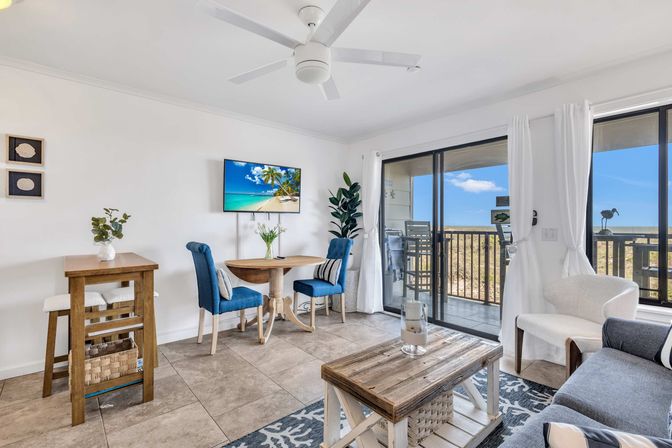 Sunny beachfront condo living room with sliding glass doors to an ocean-view balcony — white walls, ceiling fan, wooden coffee table, round dining table with blue chairs, wall TV and coastal decor.