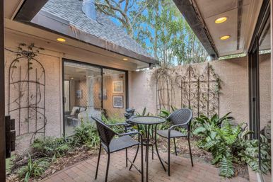 Cozy courtyard patio with round bistro table and three wicker chairs on brick pavers, leafy garden plants and wrought-iron trellises, sliding glass door to interior and recessed ceiling lights.