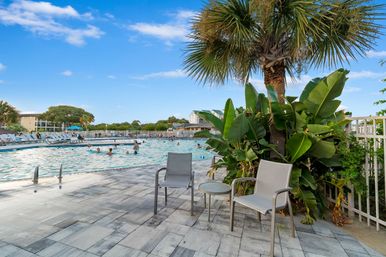 Sunny outdoor resort swimming pool with people in the water, lounge chairs and a small table on a tiled deck, framed by palm and tropical plants under a bright blue sky.