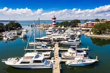 Aerial view of a sunny coastal marina with yachts and motorboats moored along wooden piers, a red-and-white striped lighthouse, waterfront buildings and calm blue water under fluffy white clouds.