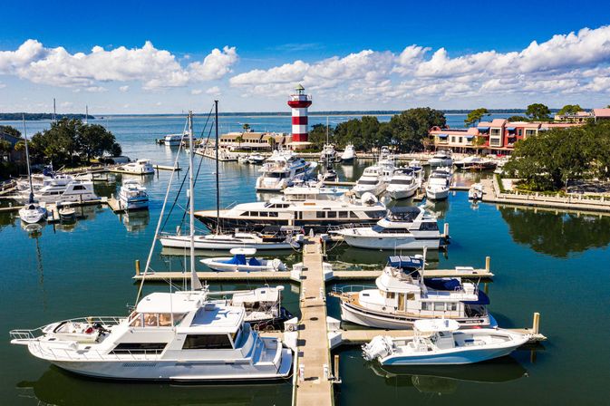 Aerial view of a sunny coastal marina with yachts and motorboats moored along wooden piers, a red-and-white striped lighthouse, waterfront buildings and calm blue water under fluffy white clouds.