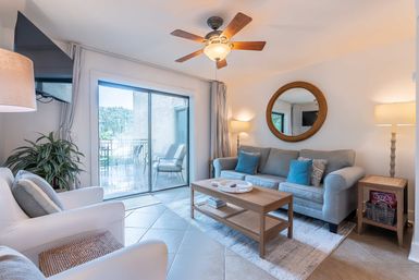 Bright, sunlit living room with gray sofa and blue accent pillows, wooden coffee table, round wall mirror, ceiling fan, tiled floor, potted plant, and sliding glass door opening to a balcony with outdoor chairs.
