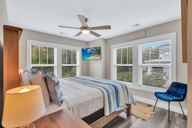 Sunlit modern coastal-style bedroom with a bed dressed in white linens and a blue-striped throw, wooden nightstand with lamp, ceiling fan, wall-mounted TV displaying a beach, four windows with white blinds, blue accent chair and hardwood floors.
