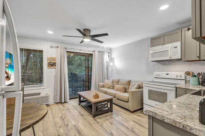 Open-concept condo living room and kitchen with beige sofa, wooden coffee table, modern ceiling fan, sliding glass door to tree-lined balcony, white stove and microwave, gray cabinets and granite countertops on light wood floors.