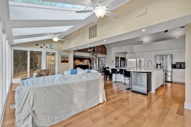 Sunlit open-concept living room and kitchen with skylights and floor-to-ceiling windows, white slipcovered sofa, hardwood floors, island with bar stools, stainless-steel appliances and spiral staircase.