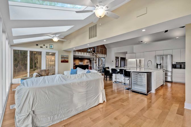 Sunlit open-concept living room and kitchen with skylights and floor-to-ceiling windows, white slipcovered sofa, hardwood floors, island with bar stools, stainless-steel appliances and spiral staircase.