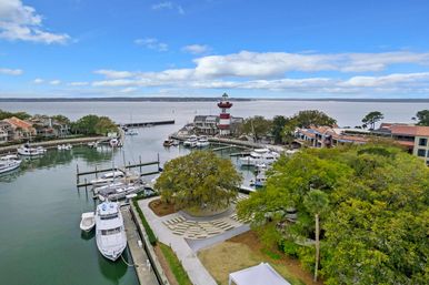 Aerial view of a coastal marina with yachts and sailboats docked around a red-and-white striped lighthouse, waterfront homes, a tree-lined park, and calm bay waters under a blue sky.