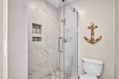 Modern coastal-themed bathroom with glass walk-in shower, white marble-look tile, built-in shower niche, mosaic floor tile, and wooden anchor wall decor above a white toilet.