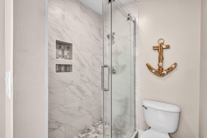 Modern coastal-themed bathroom with glass walk-in shower, white marble-look tile, built-in shower niche, mosaic floor tile, and wooden anchor wall decor above a white toilet.