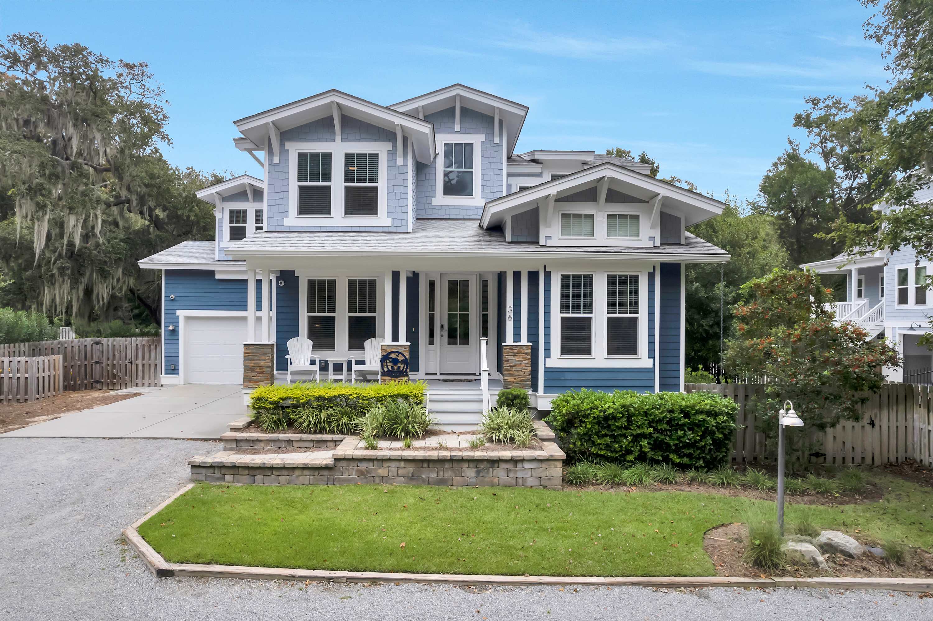 Bright blue two-story Craftsman-style home with white trim, covered front porch with chairs, attached garage, raised stone planter, manicured lawn and surrounding trees