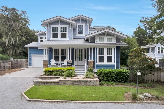 Bright blue two-story Craftsman-style home with white trim, covered front porch with chairs, attached garage, raised stone planter, manicured lawn and surrounding trees