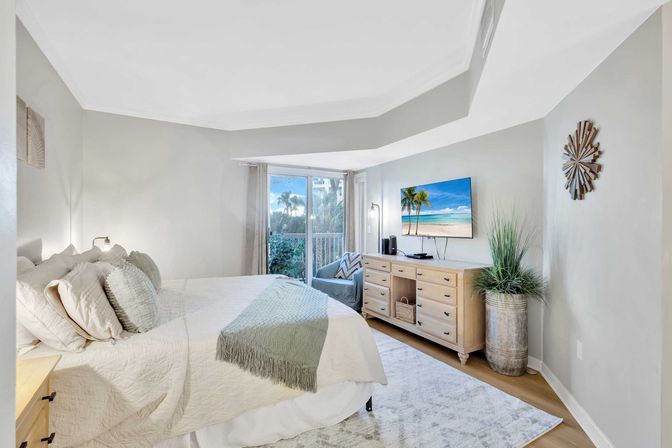 Bright, light-filled coastal bedroom with neutral linens and green throw on a queen bed, sliding glass door to a balcony with palm-tree and water glimpse, light-wood dresser with wall-mounted TV, potted plant and cozy accent chair.