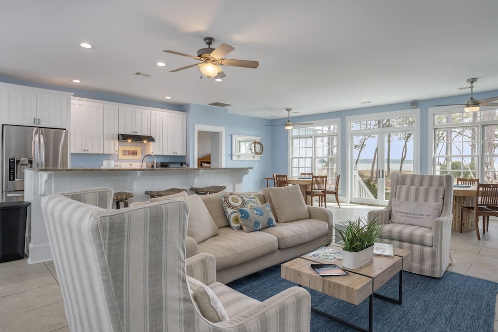 Sunlit waterfront open-plan living room and kitchen with a beige sofa, striped armchairs, wooden coffee table, blue rug, and large windows framing water and trees.