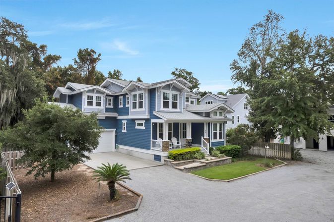 Charming blue coastal-style two-story house with covered front porch, attached garage, gravel driveway, small palm and landscaped front yard framed by mature trees