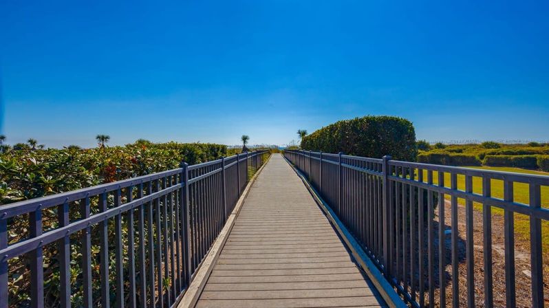 Sunny coastal wooden boardwalk with metal railings leading through green dunes to the beach horizon under a clear blue sky