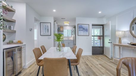 Bright open-plan dining area in a contemporary home with a white marble table, six tan leather chairs, glass vase with greenery, light wood plank flooring, built-in beverage fridge, and a view into a minimalist kitchen and entryway.
