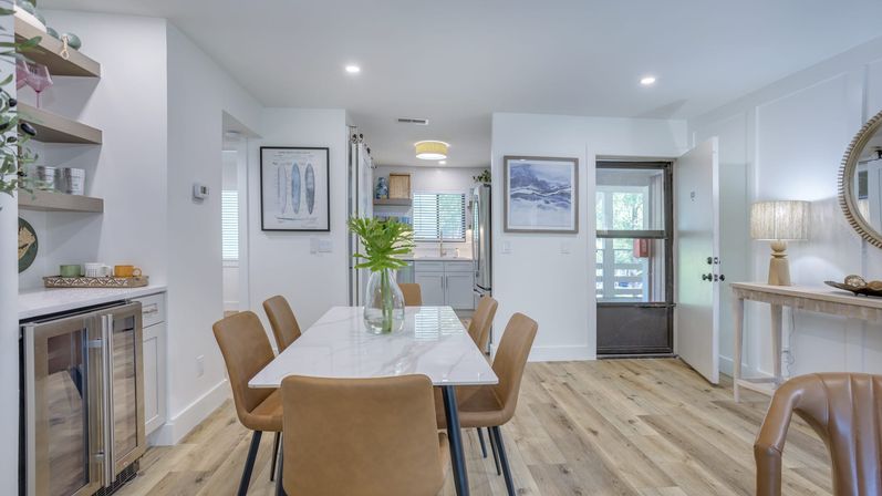 Bright open-plan dining area in a contemporary home with a white marble table, six tan leather chairs, glass vase with greenery, light wood plank flooring, built-in beverage fridge, and a view into a minimalist kitchen and entryway.