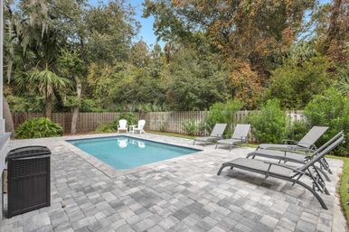 Sunny backyard swimming pool with gray paver patio, four reclining lounge chairs, two white Adirondack chairs, wooden privacy fence and dense tropical greenery