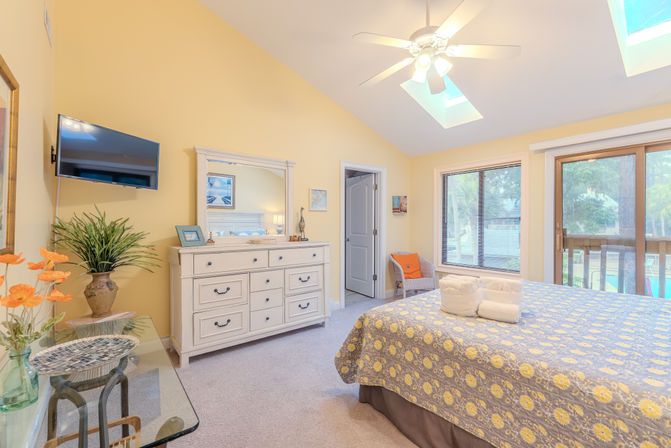 Cheerful sunny yellow bedroom with skylights and ceiling fan, white dresser with mirror, floral quilted bed with folded towels, and sliding glass door to a pool-side deck.