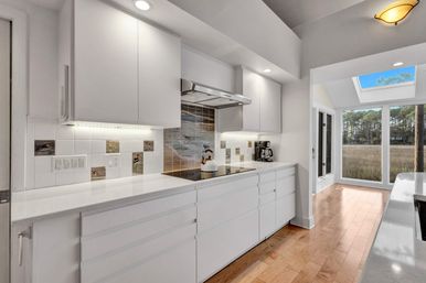 Modern white galley kitchen with quartz counters, hardwood floors, undercabinet lighting, seaside tile backsplash, skylight and floor-to-ceiling windows overlooking a coastal marsh and pine trees