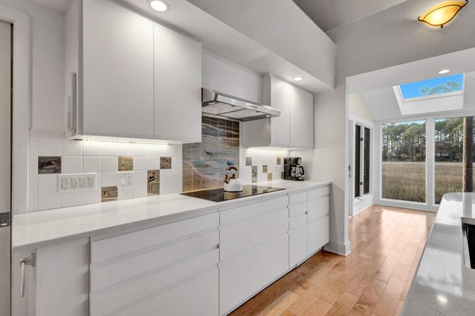 Modern white galley kitchen with quartz counters, hardwood floors, undercabinet lighting, seaside tile backsplash, skylight and floor-to-ceiling windows overlooking a coastal marsh and pine trees