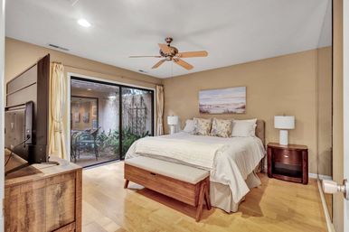 Cozy master bedroom with king bed, patterned pillows, wooden bench and dresser, ceiling fan and flat-screen TV, sliding glass doors opening to a small private patio with plants and seating.