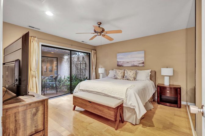 Cozy master bedroom with king bed, patterned pillows, wooden bench and dresser, ceiling fan and flat-screen TV, sliding glass doors opening to a small private patio with plants and seating.