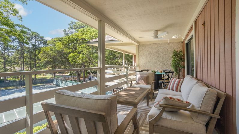 Cozy sunlit covered balcony with cushioned outdoor sofa and chairs, wooden coffee table, ceiling fan, potted plant, and striped pillows overlooking tree-lined tennis courts and greenery.