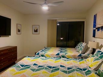 Cozy modern bedroom with two beds dressed in colorful geometric quilts, ceiling fan with light, wall-mounted TV, wooden dresser, sliding glass door with vertical blinds, bedside lamp and abstract wall art.