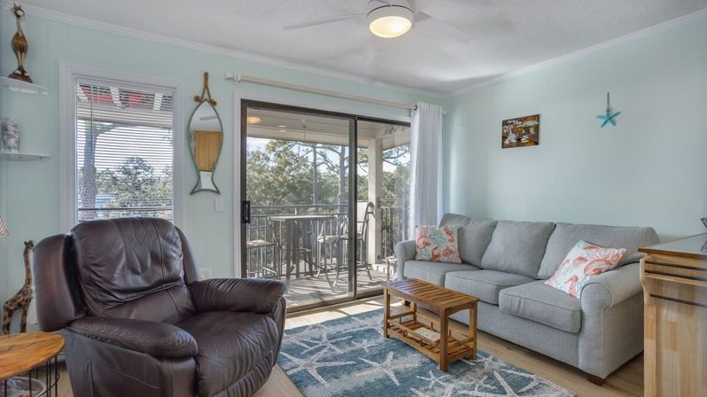 Bright coastal living room in a vacation condo with a gray sofa and coral-patterned pillows, leather recliner, wooden coffee table, blue patterned rug, ceiling fan, and sliding glass doors opening to a balcony with outdoor seating and treetop views.