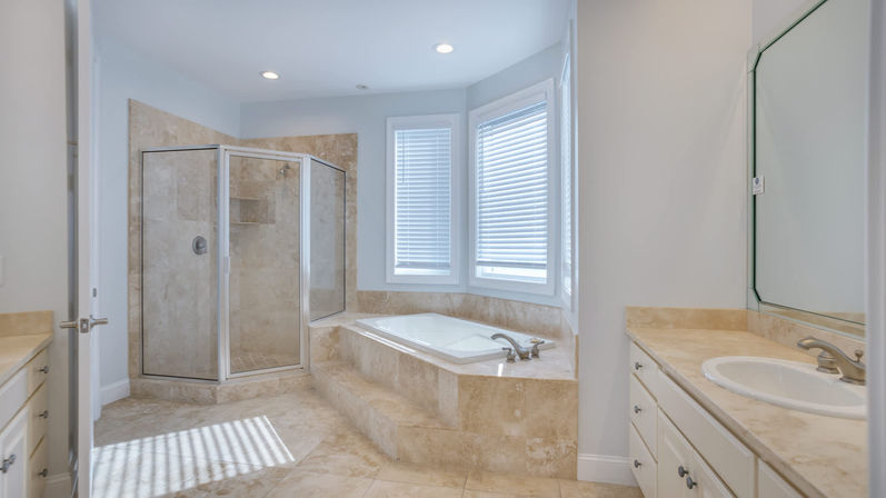Sunlit spa-like bathroom with beige marble tile, corner glass shower, and raised soaking tub beneath bay windows.