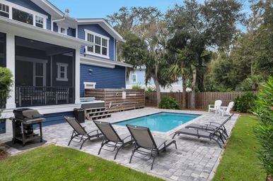 In-ground backyard pool beside a blue coastal-style house with screened porch, paver patio with reclining lounge chairs and gas grill, palm trees and wooden privacy fence.