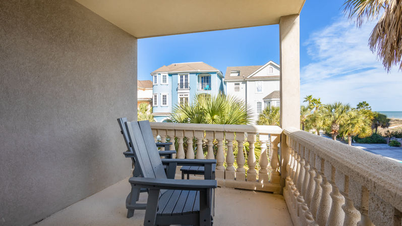 Sunny beachfront balcony with two gray Adirondack chairs, balustrade railing, palm trees, pastel coastal homes and ocean horizon under a bright blue sky.