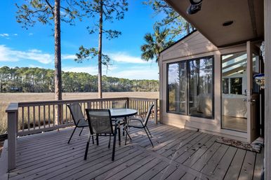 Sunlit wooden marshfront deck with round glass-top patio table and four mesh chairs, overlooking coastal marsh grasses and pine trees with sliding glass doors to the home.