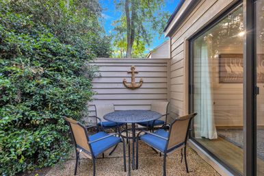 Cozy private courtyard patio with round metal dining table, four blue‑cushioned chairs, anchor wall decor, sliding glass door and lush greenery.