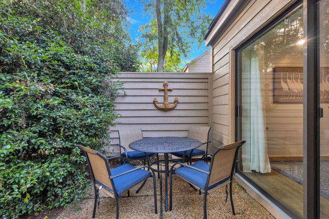 Cozy private courtyard patio with round metal dining table, four blue‑cushioned chairs, anchor wall decor, sliding glass door and lush greenery.
