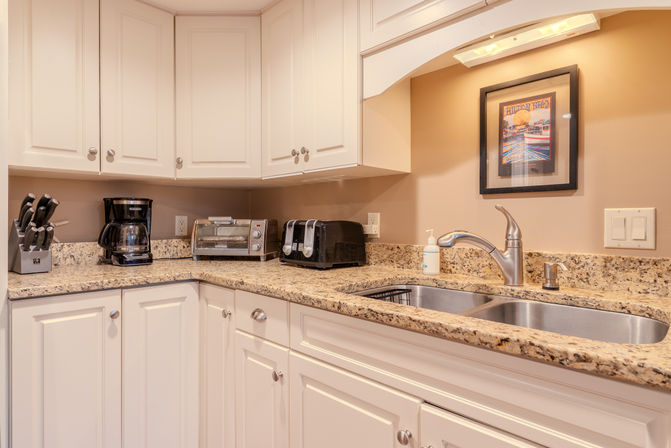 Bright modern kitchen corner with white cabinets, speckled granite countertop, stainless double sink and chrome faucet, black coffee maker, toaster, toaster oven and knife block under warm under-cabinet lighting.