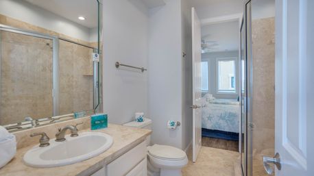 Bright ensuite bathroom with beige tiled walk-in shower, marble vanity and sink, toilet, and open door leading to a bedroom with blue starfish-patterned bedding and windows.