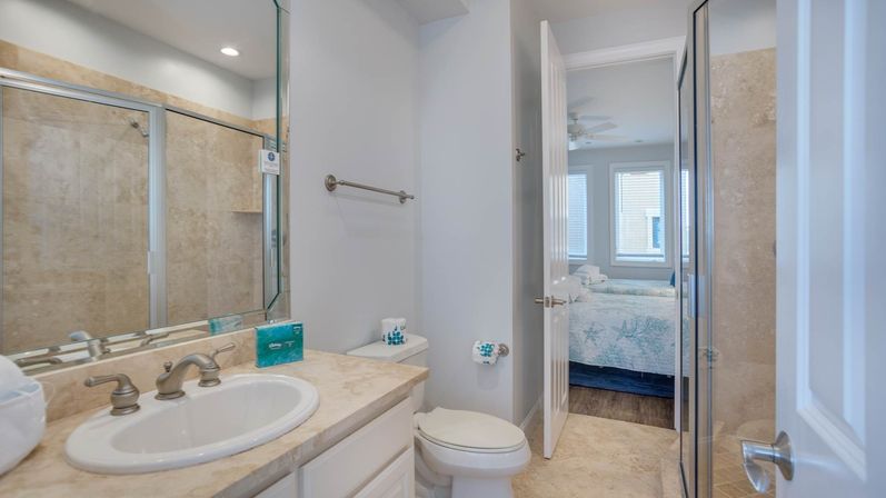 Bright ensuite bathroom with beige tiled walk-in shower, marble vanity and sink, toilet, and open door leading to a bedroom with blue starfish-patterned bedding and windows.