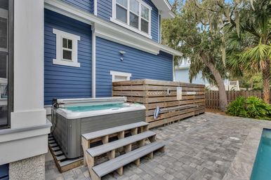 Backyard hot tub with wooden steps beside a blue coastal-style house, slatted privacy fence, paver patio, palm tree and pool edge visible