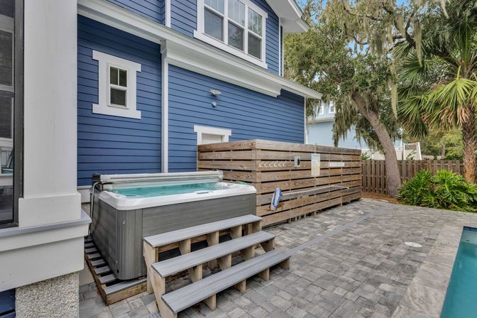 Backyard hot tub with wooden steps beside a blue coastal-style house, slatted privacy fence, paver patio, palm tree and pool edge visible