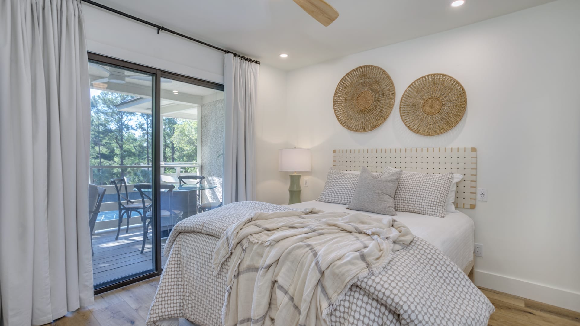 Sunlit neutral bedroom with textured bedding, woven wall baskets and sliding glass door opening to a balcony with outdoor seating and tree view