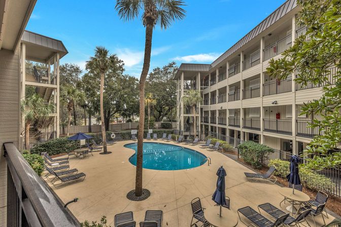 Kidney-shaped outdoor pool with lounge chairs, blue umbrellas and palm trees in a courtyard surrounded by a four-story hotel-style building with balcony walkways.