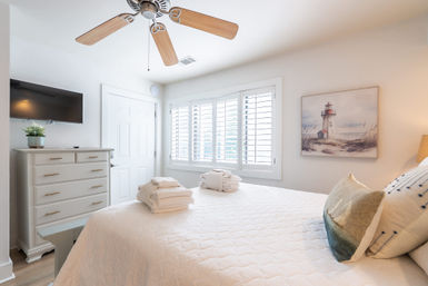 Sunlit coastal-inspired bedroom interior with white quilted bed topped with folded towels, plantation shutters, wooden-blade ceiling fan, dresser with wall-mounted TV, and lighthouse wall art.