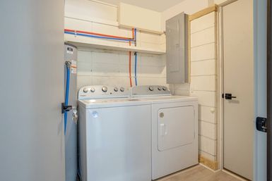 Compact basement laundry room with white top-load washer and matching dryer, visible water heater, exposed red and blue supply lines and electrical panel
