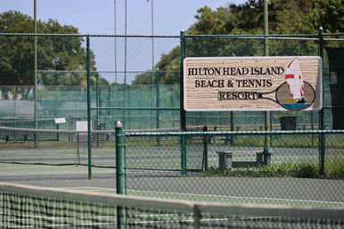 Fenced outdoor tennis courts at a sunny coastal island beach resort, wooden sign with a sailboat graphic