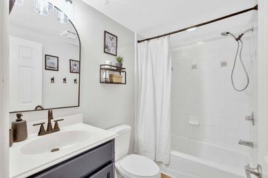 Bright modern bathroom with white tub-shower and textured curtain, oil-rubbed bronze shower and faucet, dark vanity with integrated sink, arched mirror and floating shelf with plant and basket