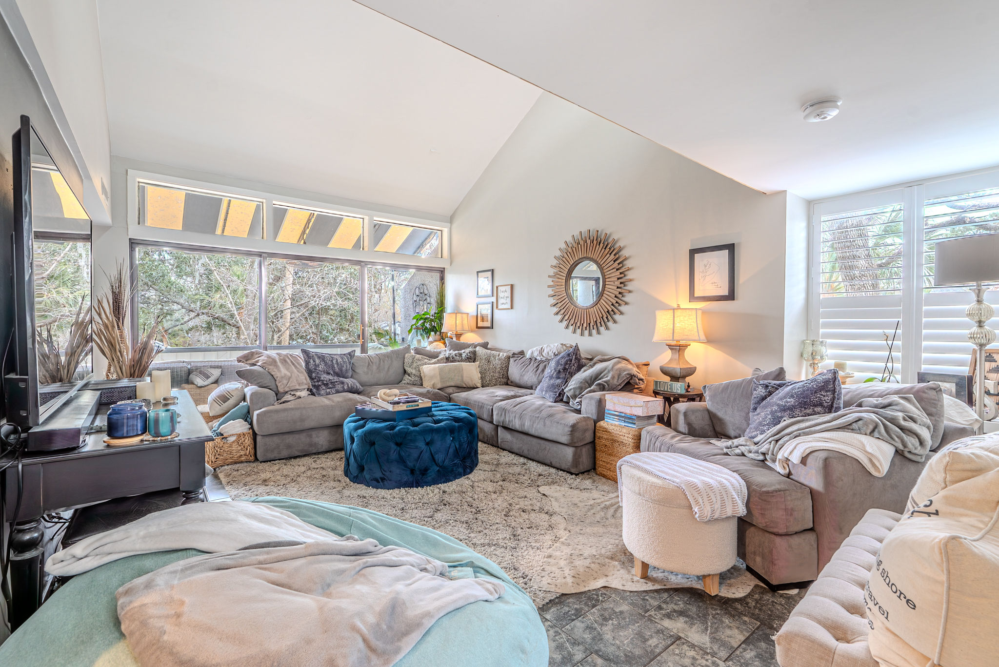 Sunlit cozy living room with oversized gray sectional, tufted blue ottoman, shag rug, sunburst mirror and large windows overlooking trees